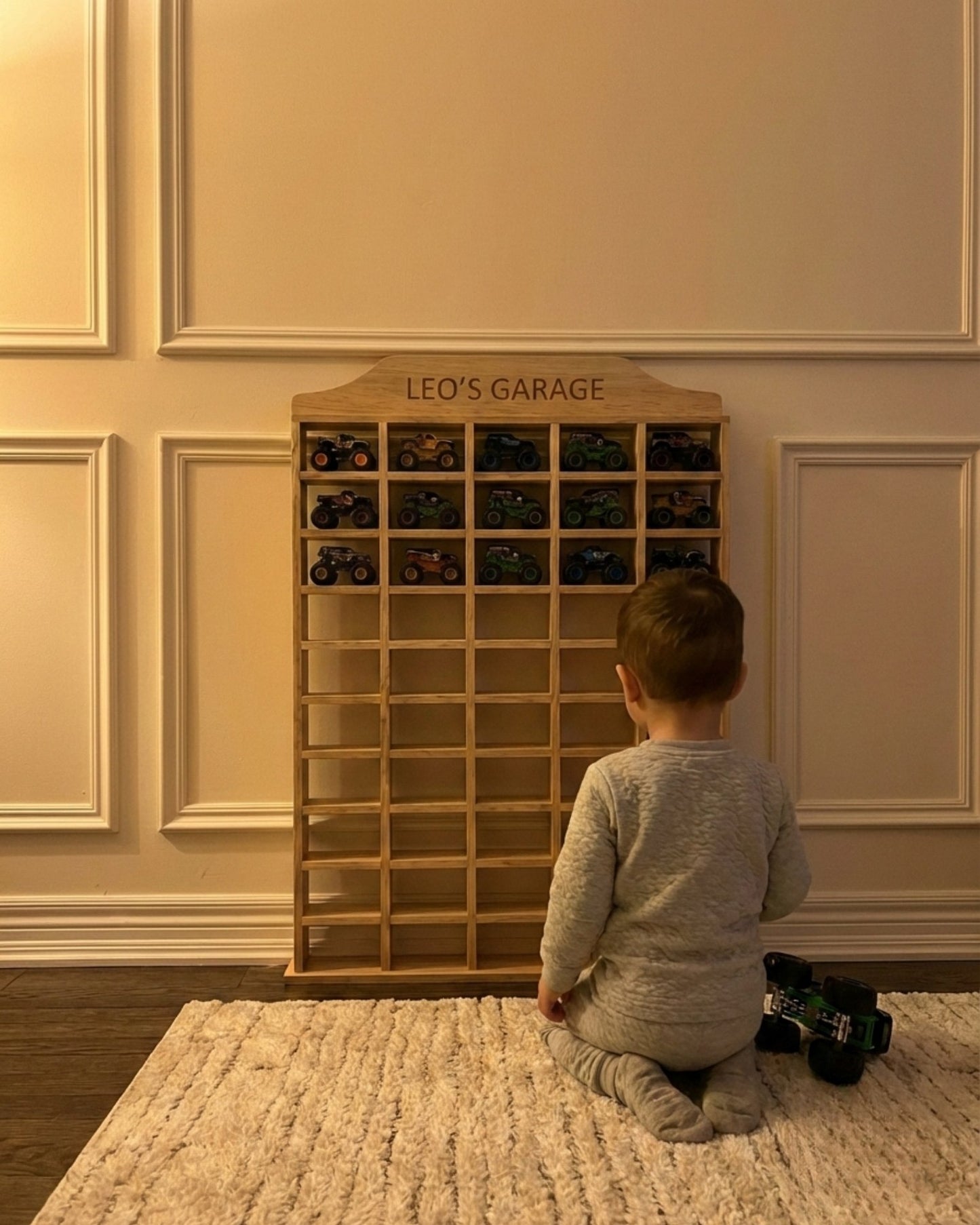 Child looking at a wooden toy car organizer with 'Leo's Garage' on a carpeted floor.