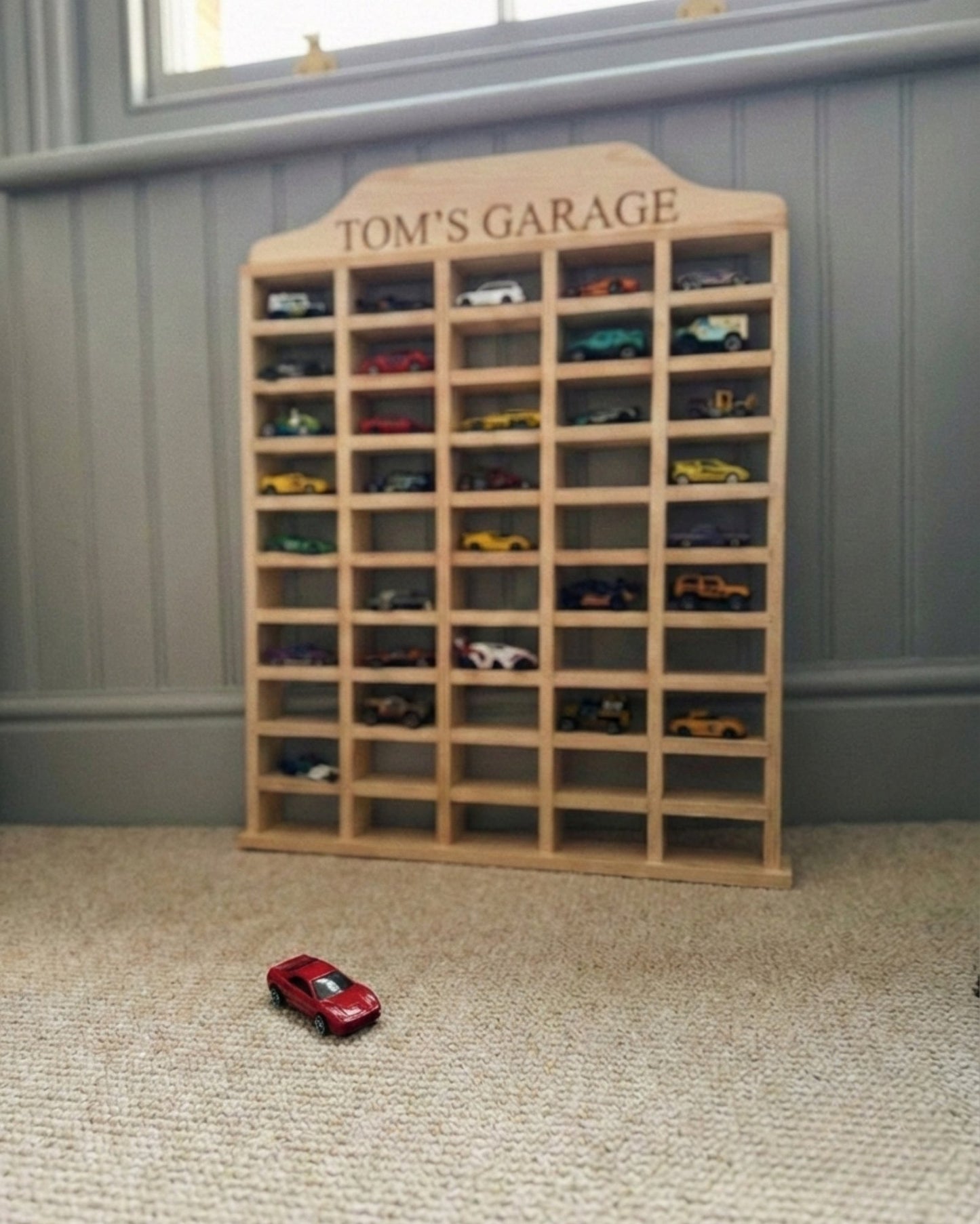 Wooden model car display case with 'Tom's Garage' on top, filled with model cars, in a room with a gray wall.
