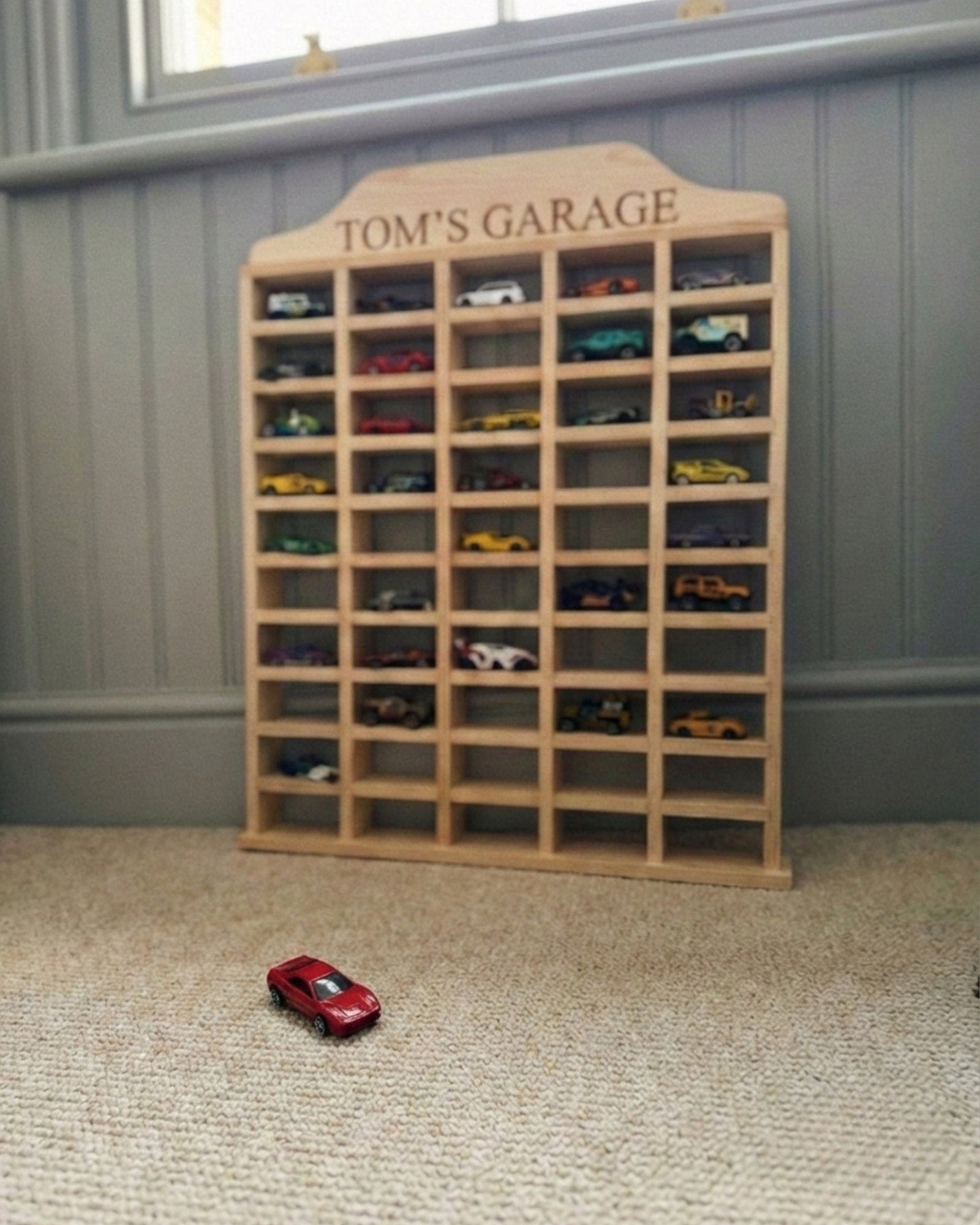 Wooden model car display case with 'Tom's Garage' on top, filled with model cars, in a room with a gray wall.