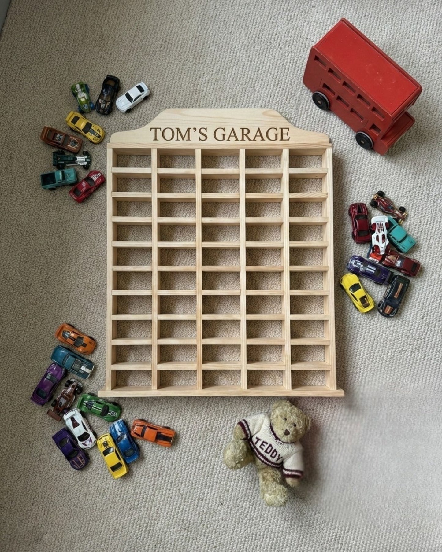 Wooden toy car garage with 'Tom's Garage' branding, surrounded by miniature cars and a teddy bear on a carpeted floor.
