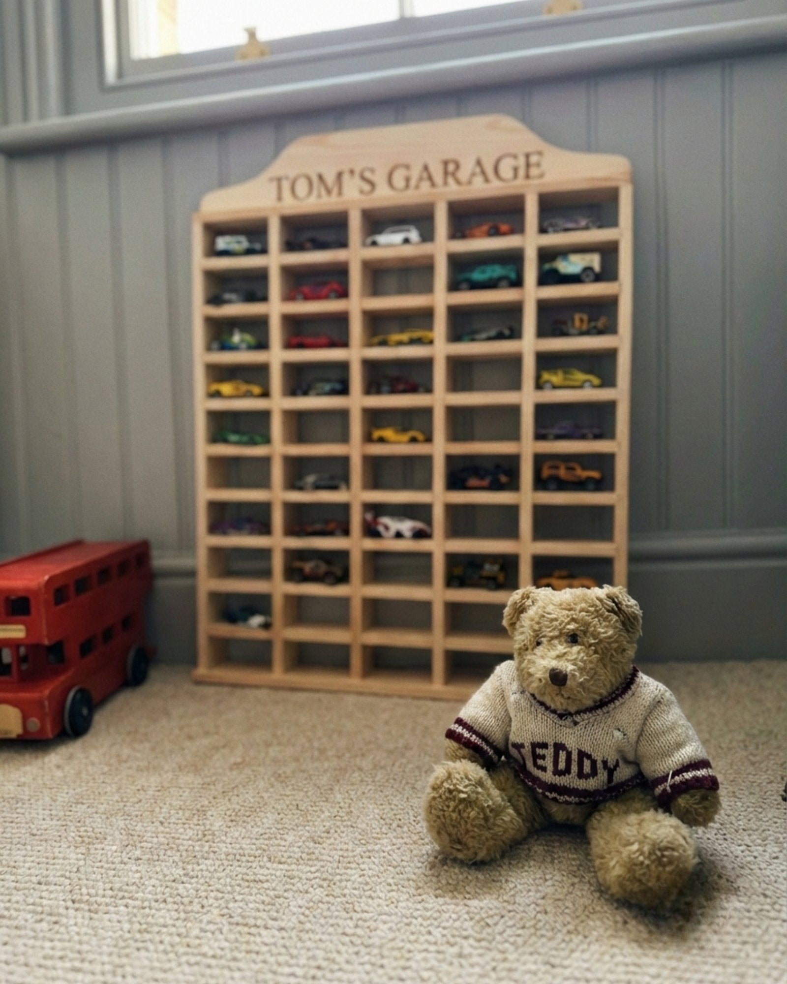 Wooden toy car display case with 'Tom's Garage' branding, featuring various model cars, and a teddy bear sitting in front.