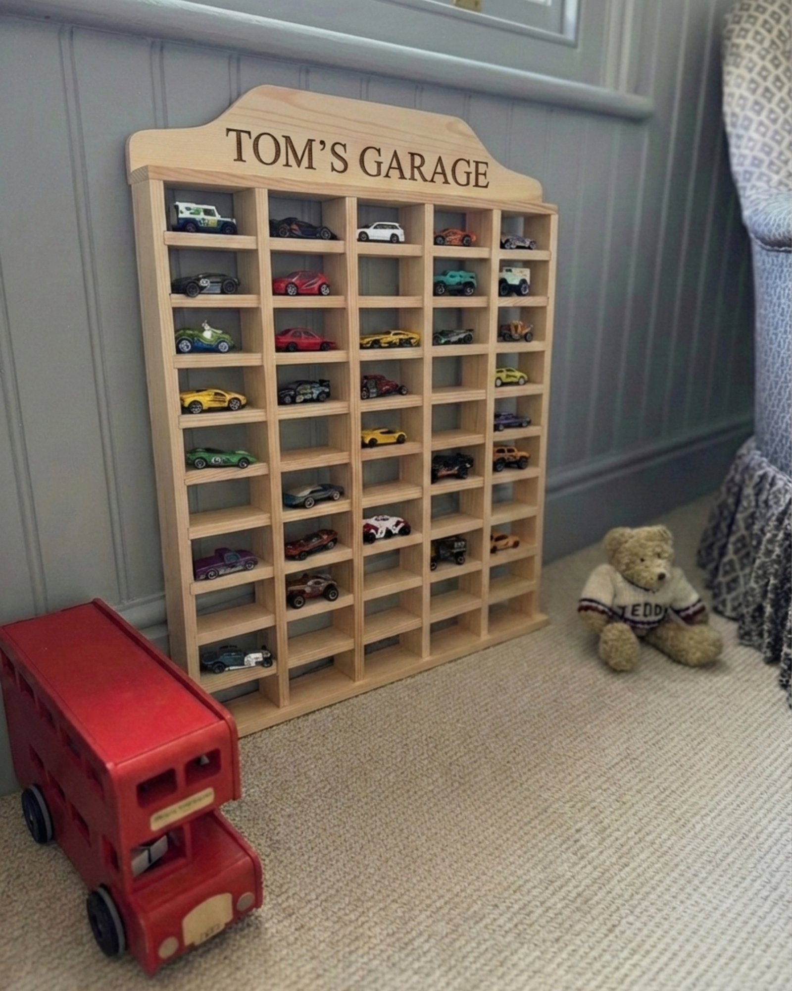 Wooden toy car garage with 'Tom's Garage' on top, filled with toy cars, next to a red toy bus and teddy bear.