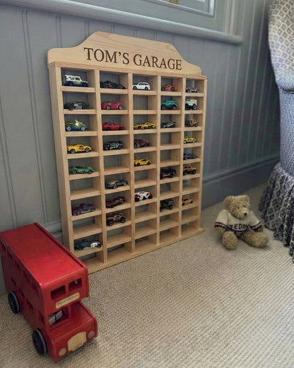 Wooden toy car garage with 'Tom's Garage' on top, filled with toy cars, next to a red toy bus and teddy bear.